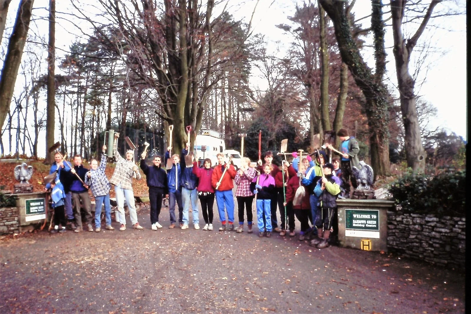 Group of children and adults standing outdoors in a forested area holding shovels, some leaning on or raising shovels, in front of a sign that says "Welcome to Baileyton Gibbon Playground".