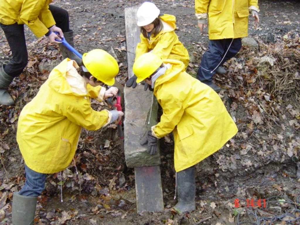 Children and adults in yellow raincoats and helmets working together to install a concrete marker or boundary stone in a muddy outdoor area.