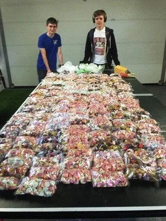 Two teenage boys standing behind a large table filled with hundreds of zippered plastic bags containing toy or candy-like items, in an indoor setting.