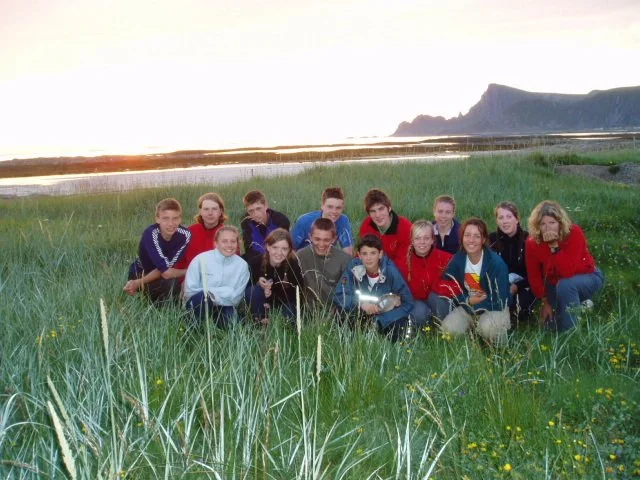 Group of people sitting on grass near a beach with water and cliffs in the background during sunset.