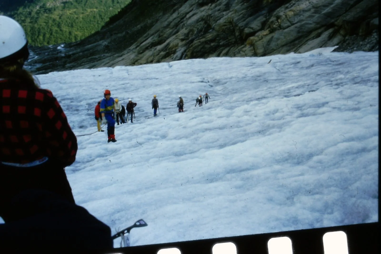 A group of people wearing helmets and outdoor gear trekking across a snowy and icy mountain slope, with a person in the foreground observing or participating.