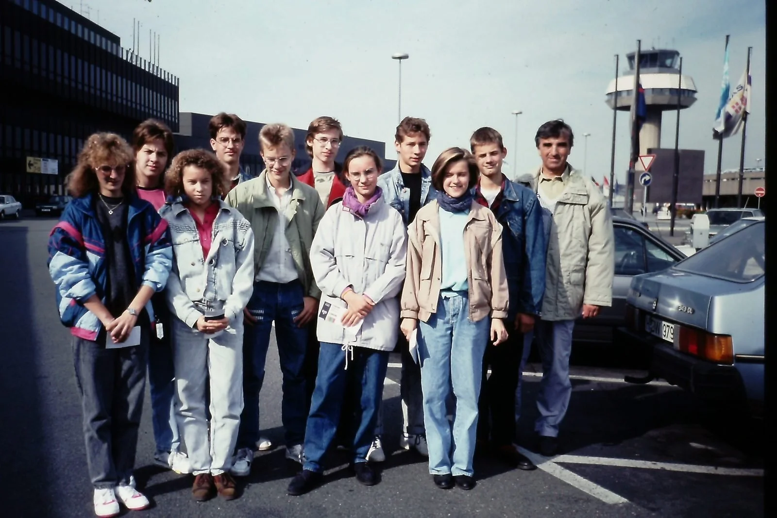 A group of eleven teenagers and one adult standing in a parking lot, with parked cars and an airport control tower in the background.