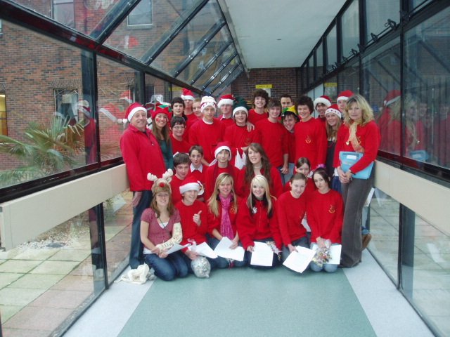 Group of children and adults wearing red Christmas sweaters and Santa hats, posing for a group photo in a glass-enclosed walkway.