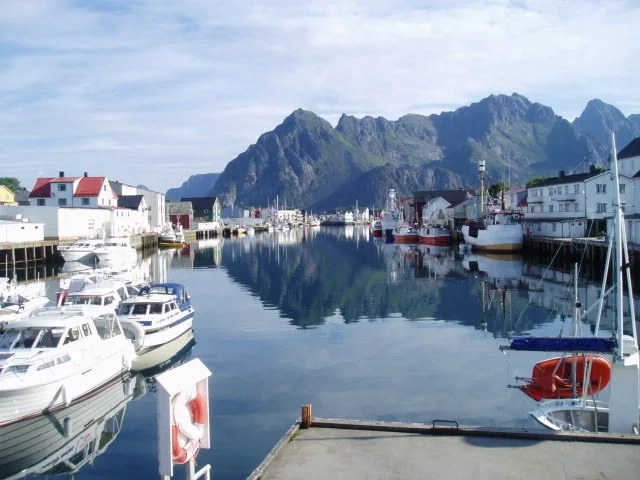 Harbor with boats docked along the water, mountains in the background, and buildings lining the shores.