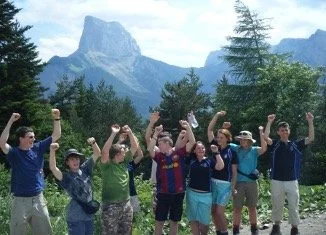 Group of children and teenagers outdoors with mountains and trees in the background, raising their hands in celebration.