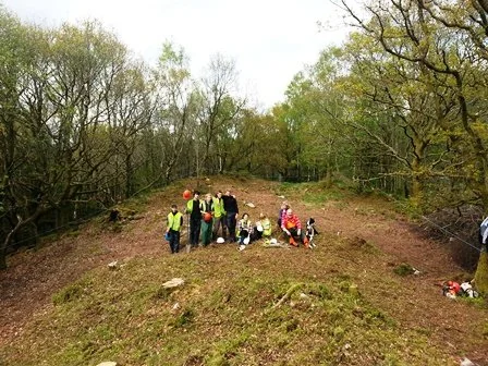 Group of people outdoors on a grassy hill, surrounded by trees, some sitting and some standing