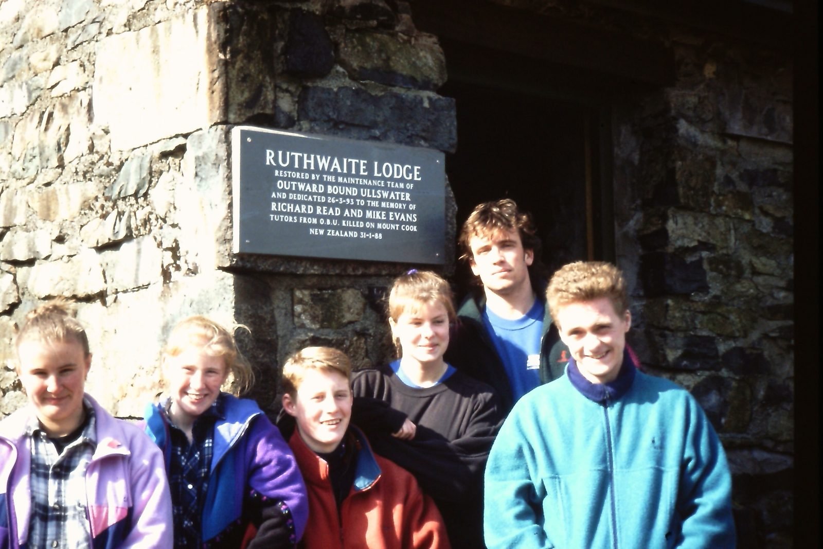 Group of six young people standing in front of a stone wall with a plaque, smiling at the camera outdoors.