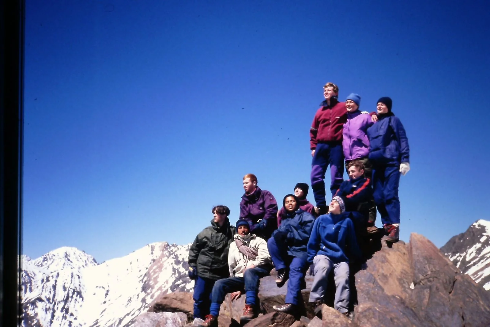 A group of hikers, including children and adults, standing on a rocky mountain peak with snow-covered mountains in the background under a clear blue sky.
