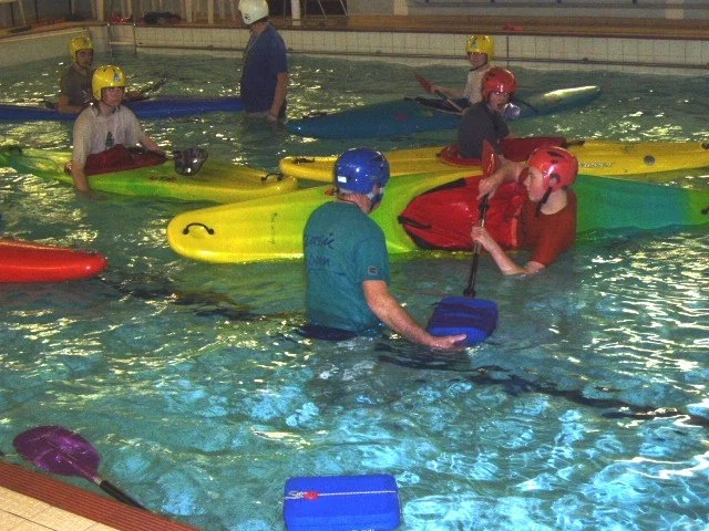 Group of people participating in a kayaking class in an indoor swimming pool, wearing helmets and life jackets, with some instructors aiding students.