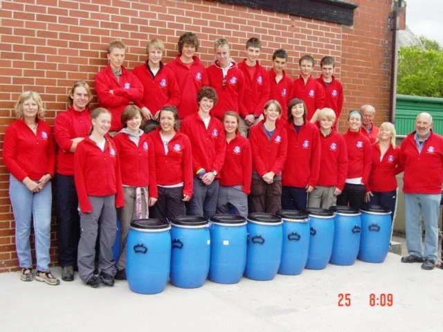 Group of people standing outdoors in front of a brick wall, wearing matching red jackets, with blue barrels on the ground in front of them.