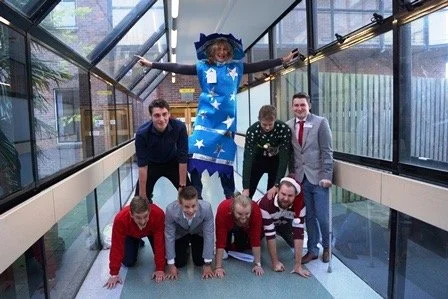 Group of people performing a human pyramid with a person dressed as a wizard on top, inside a glass corridor.