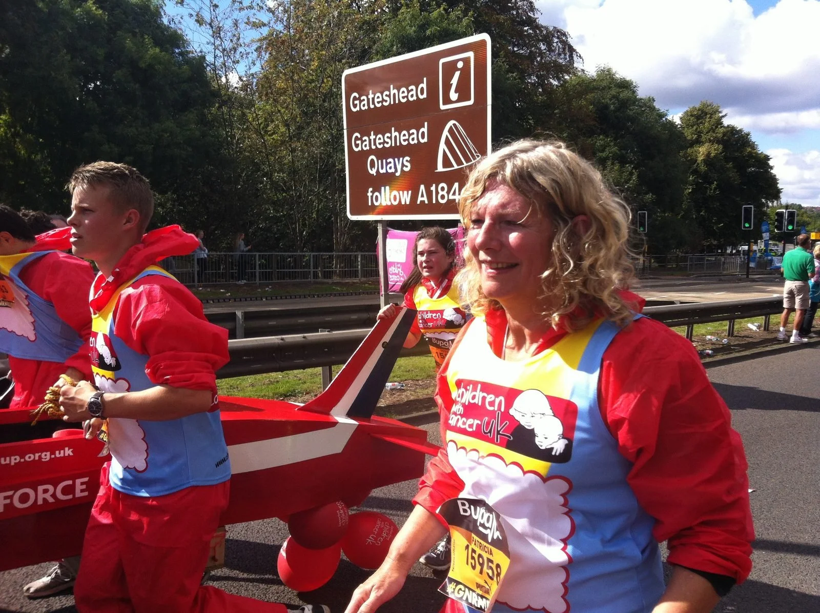People participating in a charity event wearing colorful shirts, with a woman smiling in the foreground, standing near a signpost on a cloudy day.