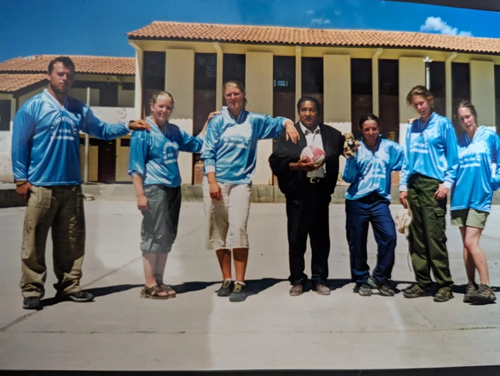 Group of six young women and one man standing outdoors in front of a building, all wearing matching light blue jerseys, with one man holding a rugby ball and a stuffed toy.