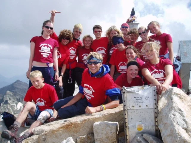 Group of people wearing red shirts on a mountain summit, celebrating with some raising their hands, with a rock formation and cloudy sky in the background.
