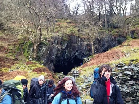 Group of hikers walking near a rocky cave entrance surrounded by trees.