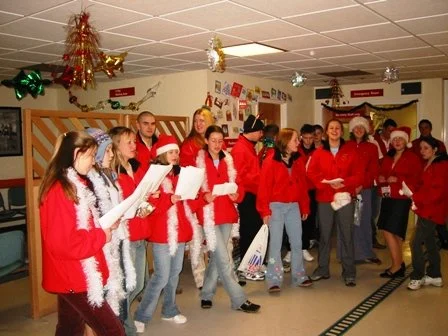 Group of people dressed in Christmas sweaters and Santa hats singing or performing during a holiday celebration indoors, decorated with Christmas ornaments and festive decorations.