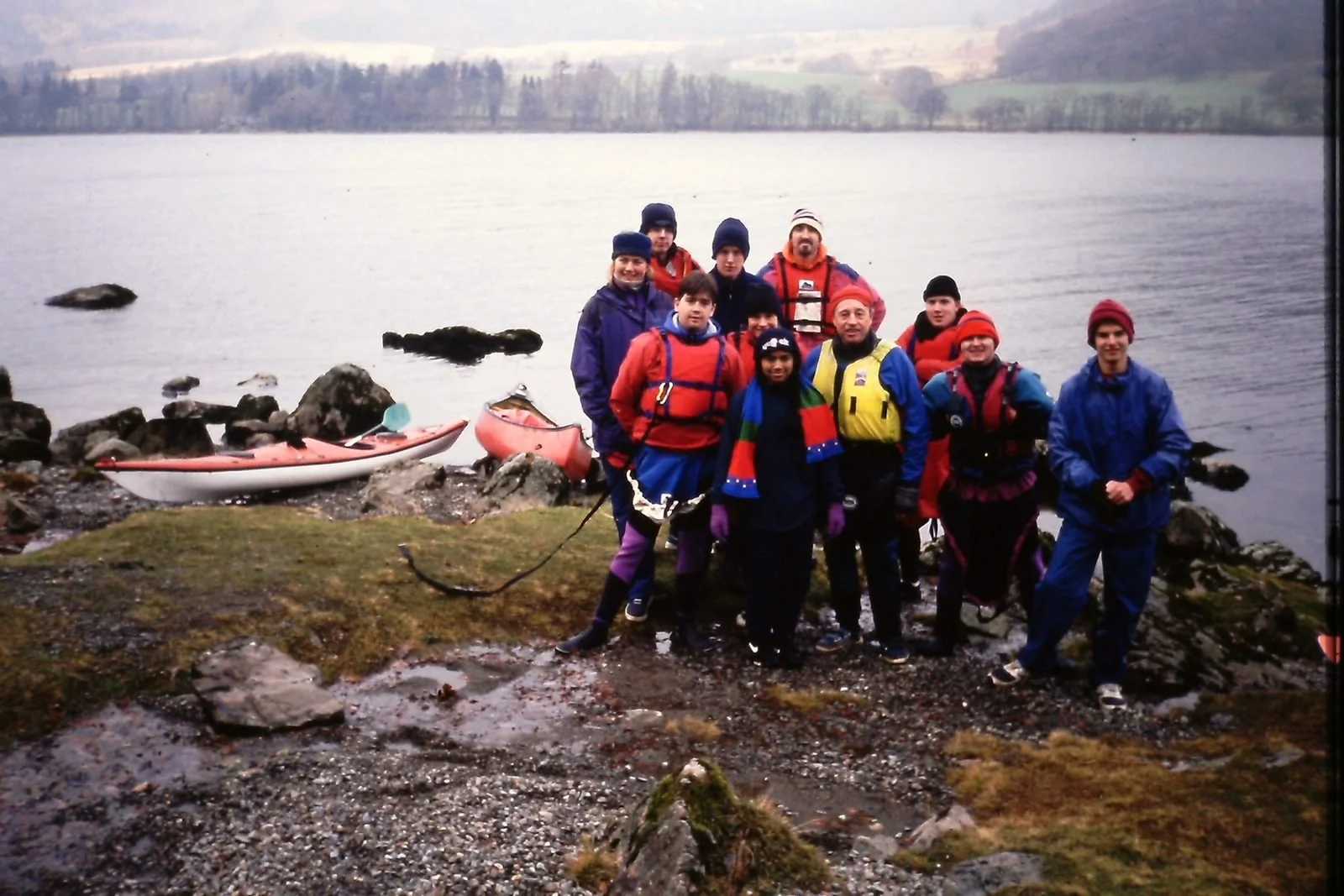 Group of people dressed in waterproof outdoor clothing standing on rocky shoreline near lake, with kayaks in the background and trees in the distance.