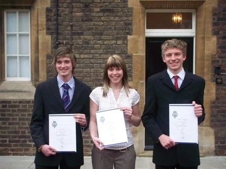 Three young people standing outside a brick building, holding certificates and smiling at the camera.