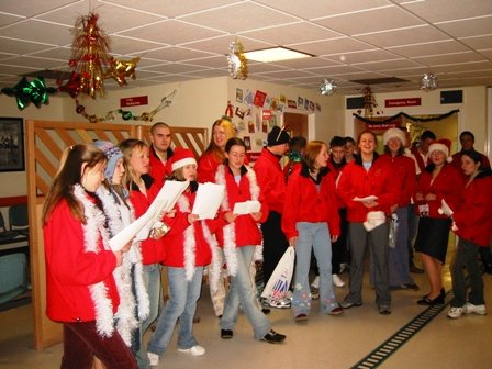 A group of young people dressed in Christmas attire, singing or performing in a decorated room with holiday decorations hanging from the ceiling.