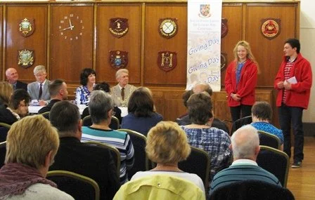 A group of people gathered in a room with wood-paneled walls and various coat of arms on the wall. Two individuals, a woman and a man, are standing in front of a large vertical banner that says "Giving Day" and "Giving Day" again. The audience is sit