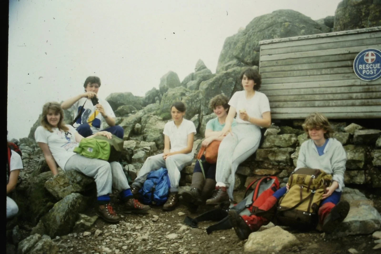Group of six people sitting on rocks and gravel near a rescue post sign, with a wooden shelter behind them, on a foggy rocky hillside, some holding backpacks and snacks.