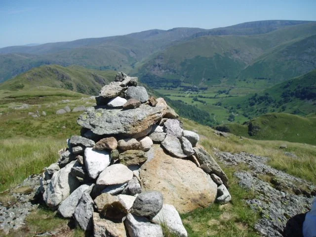 A pile of rocks on a grassy hillside in a green valley surrounded by mountains under a clear blue sky.