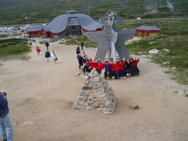 Group of children in red jackets posing around a large, abstract stone sculpture resembling a bird in an outdoor area with a building and green hills in the background.