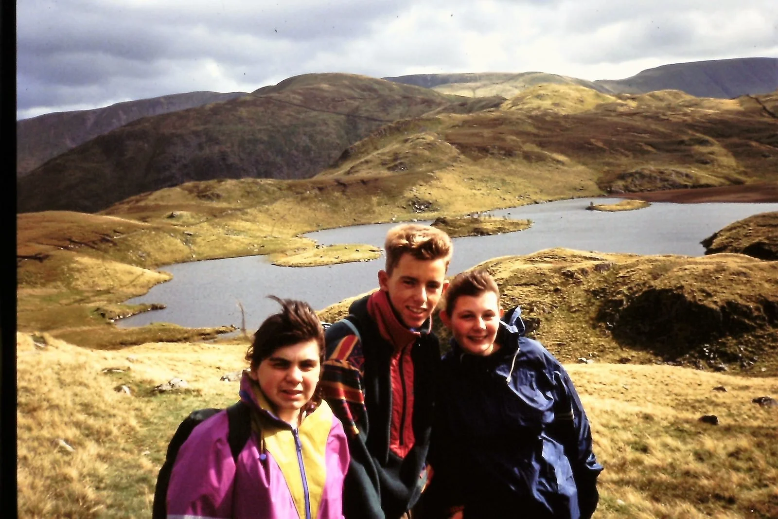 Three friends smiling and hiking near a lake in a mountainous landscape with green hills and cloudy sky.