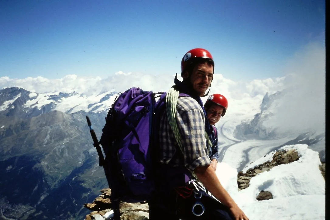 Two male climbers with helmets and backpacks sitting on a snowy mountain peak with a glacier and mountain range in the background.