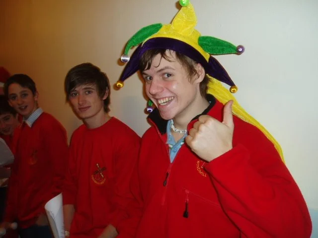 A person wearing a colorful jester hat and a red jacket giving a thumbs up, with three other people in red jackets in the background.