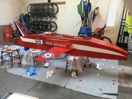 A large model of a Royal Air Force jet plane in red and white colors inside a workshop, with tools and equipment in the background.
