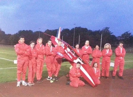 Group of people in red uniforms standing on a field with model airplane.