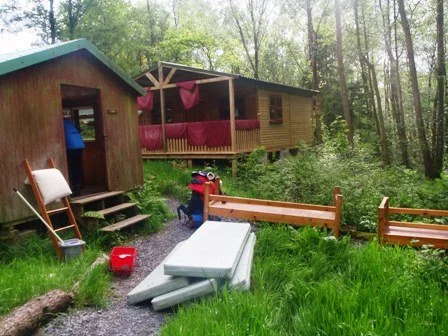Two wooden cabins in a wooded area with a small outdoor playground, containing a slide, benches, a red cooler, and scattered outdoor toys.