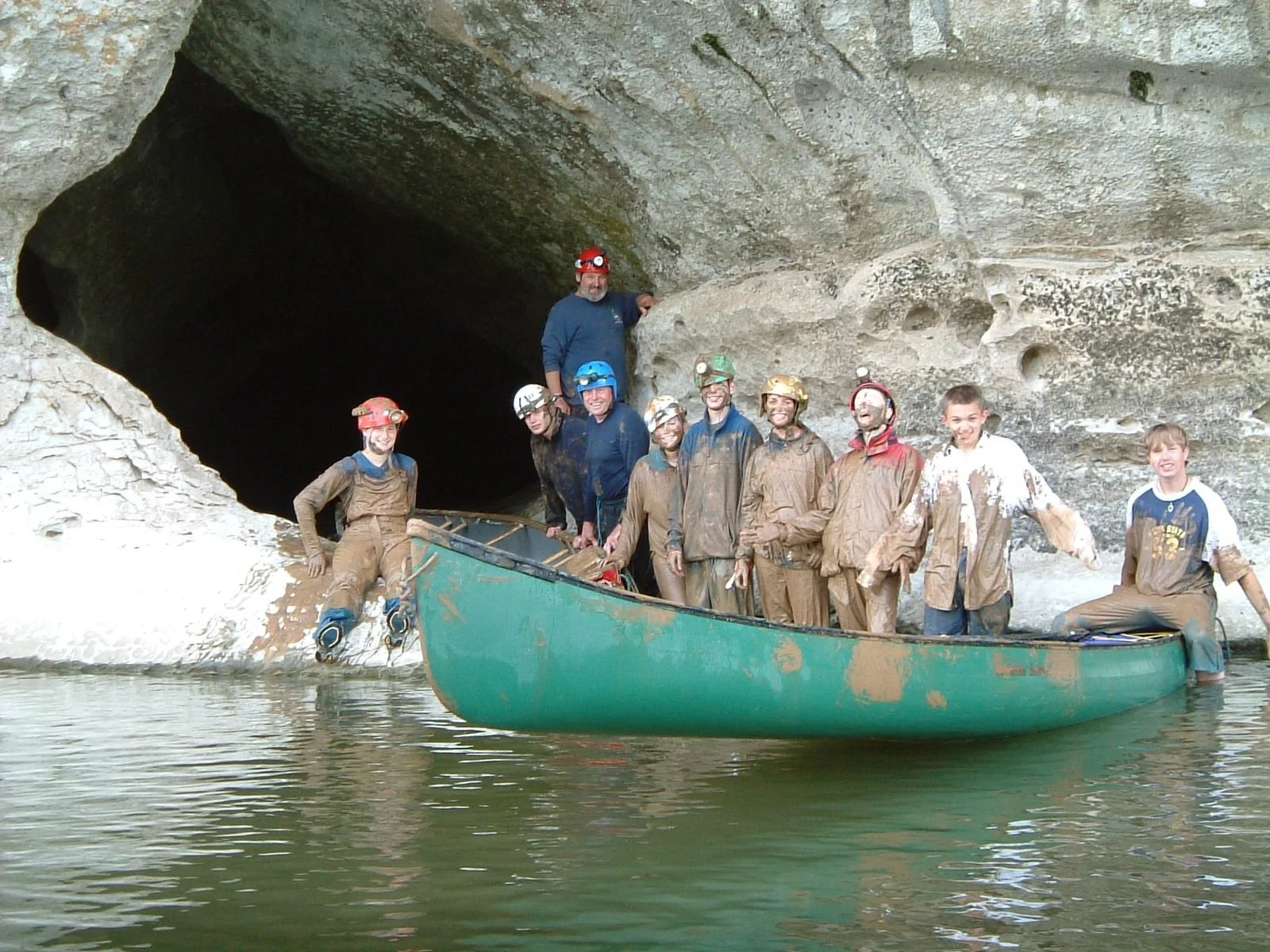 A group of people in a boat and on the rocks near a cave entrance, wearing muddy clothes and helmets after a caving or rafting adventure.