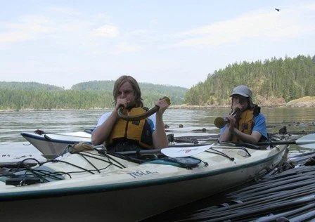 Two children in a kayak on a lake, each playing a small wind instrument, with a forested shoreline in the background.
