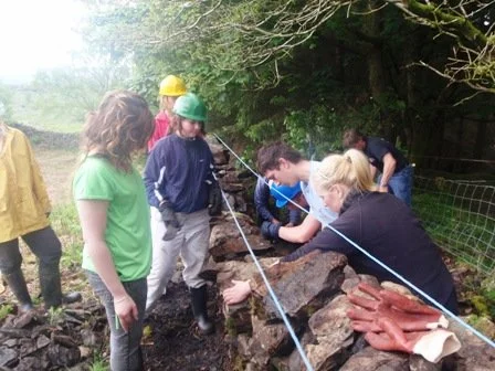Group of children and an adult observing a rock formation outdoors, with some wearing helmets and waterproof boots, on a rainy day.