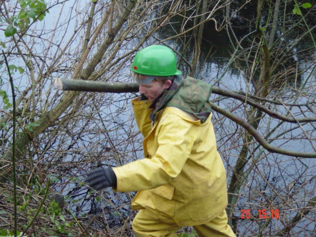 Person wearing a green helmet, yellow waterproof suit, and black gloves navigating through dense, tangled reeds and branches near a waterway.