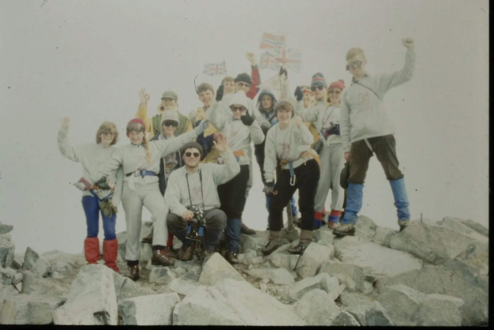A group of people in outdoor winter gear on a rocky mountain summit, holding Union Jack flags, celebrating their achievement.