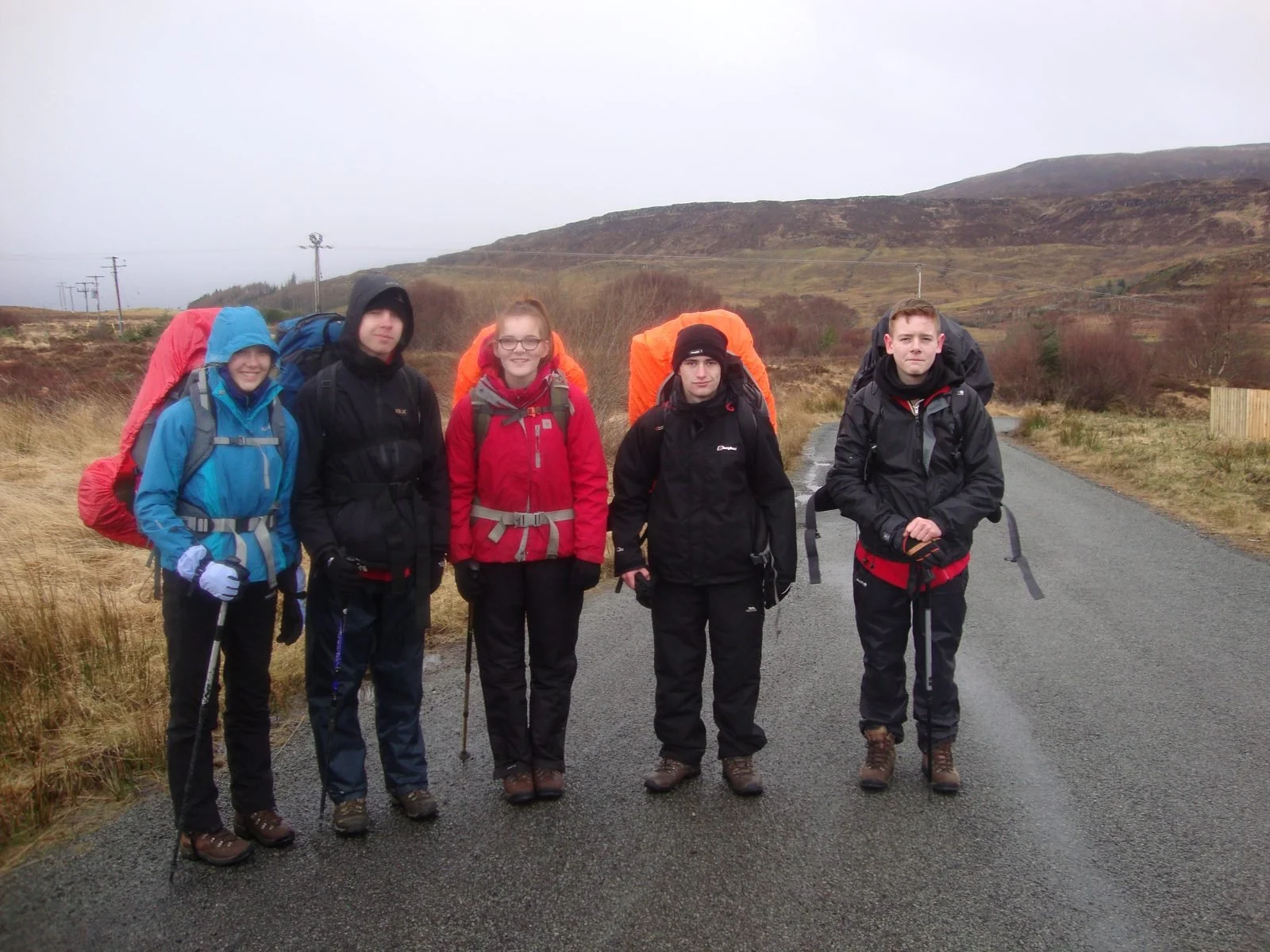 Five hikers dressed in waterproof jackets and gear standing on a rural road in a hilly, grassy landscape on a cloudy day.