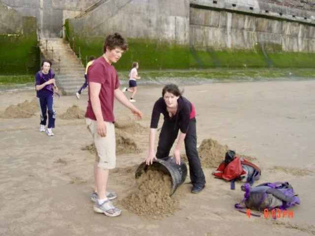 People at the beach building sandcastles, with a woman pouring sand from a bucket, and backpacks on the ground.