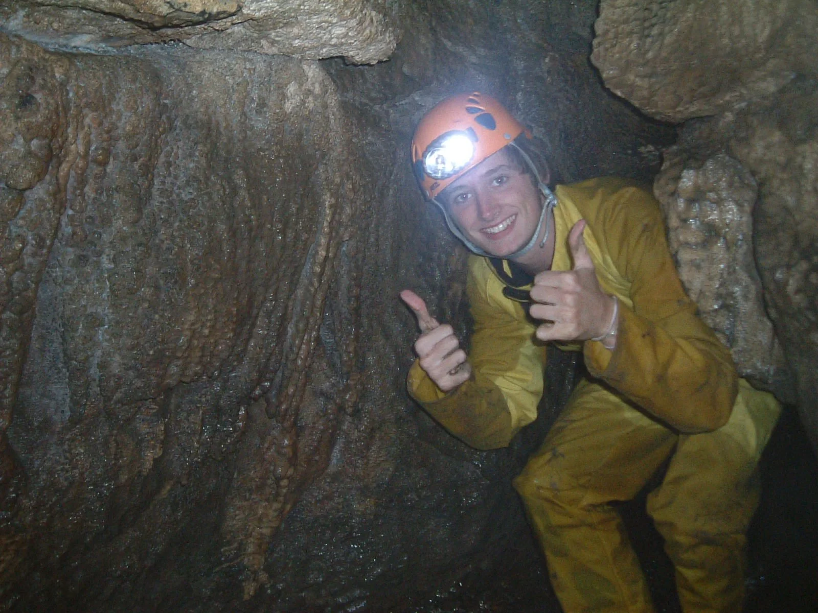 A man in yellow caving gear with a helmet giving a thumbs up while crawling inside a wet cave.