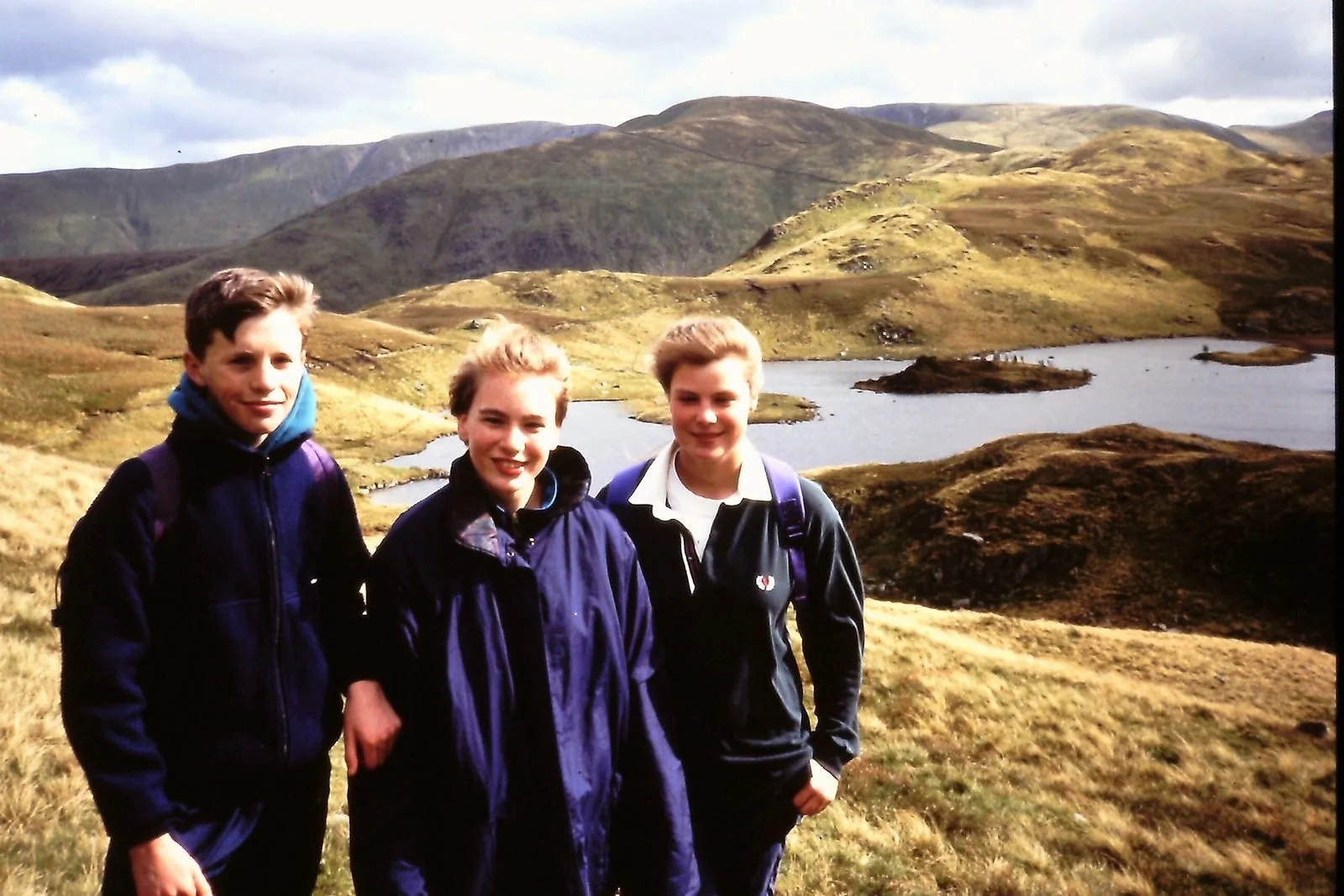 Three teenagers standing outdoors in a grassy landscape with rolling hills, lakes, and mountains in the background.