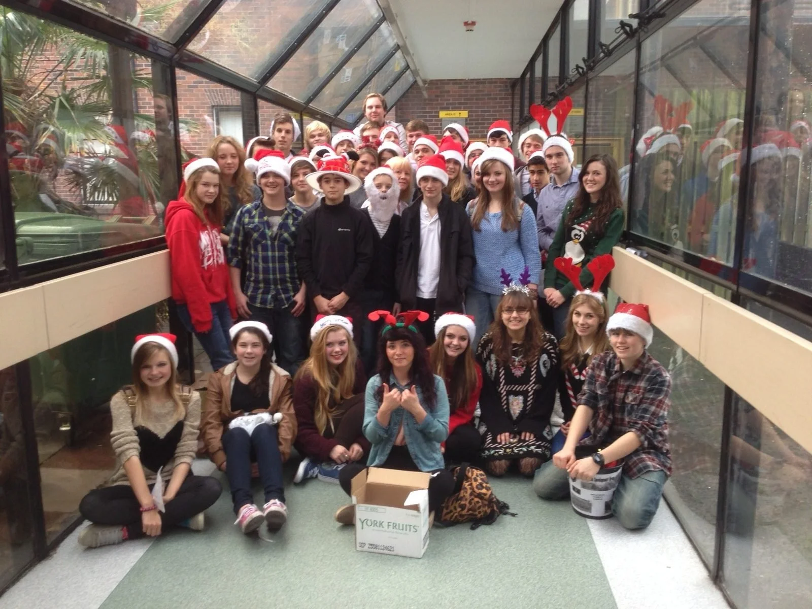 A group of young people dressed in Christmas-themed hats and accessories posing in a glass-enclosed walkway with brick building and greenery outside.