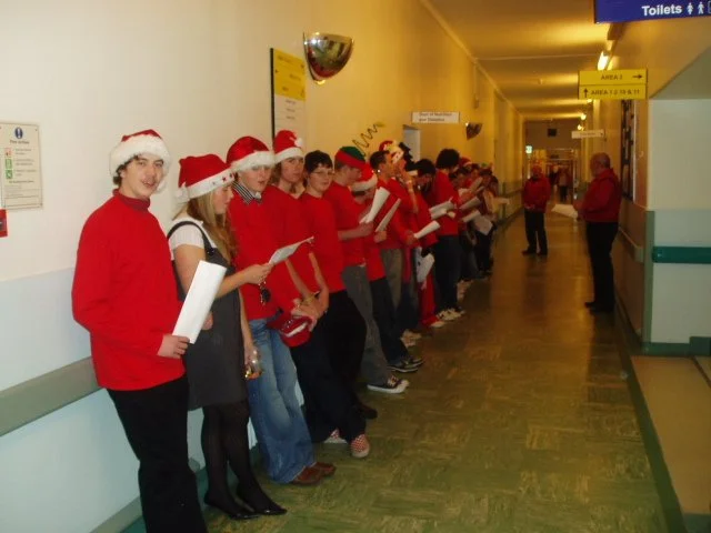 A group of people wearing Santa hats and red shirts lining up in a hospital hallway, holding papers, possibly waiting for a holiday event.