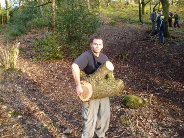 A man is holding a large cut tree trunk in a forested area, with other people visible in the background, some of whom are wearing helmets and appear to be hiking or exploring.