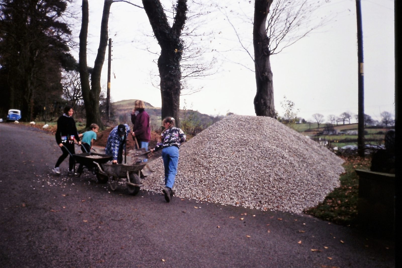 People working with shovels and wheelbarrows on a gravel pile on a rural road surrounded by trees and open fields.