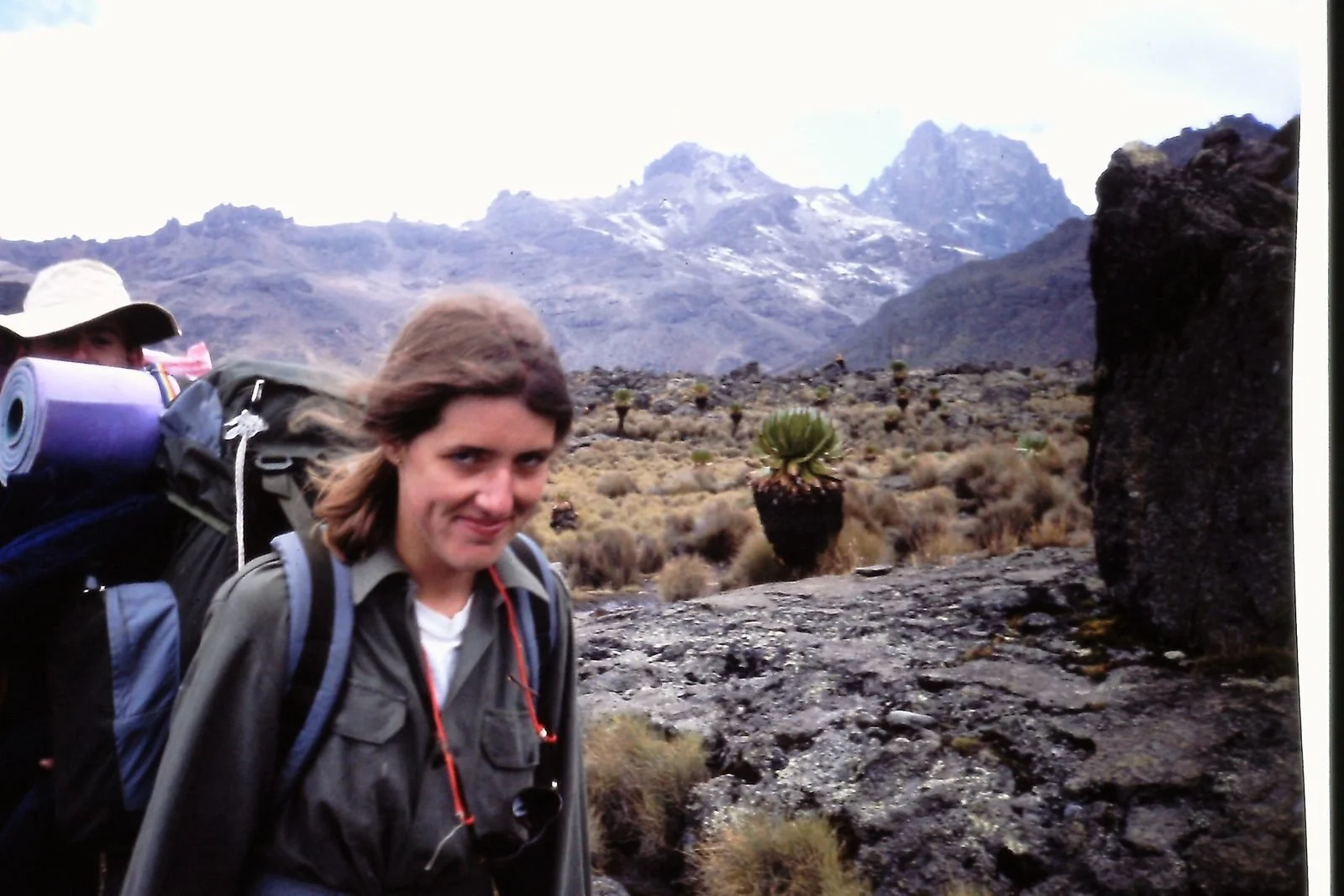 Two hikers in a mountainous desert landscape with sparse vegetation, cacti, and rocky terrain, under cloudy skies.