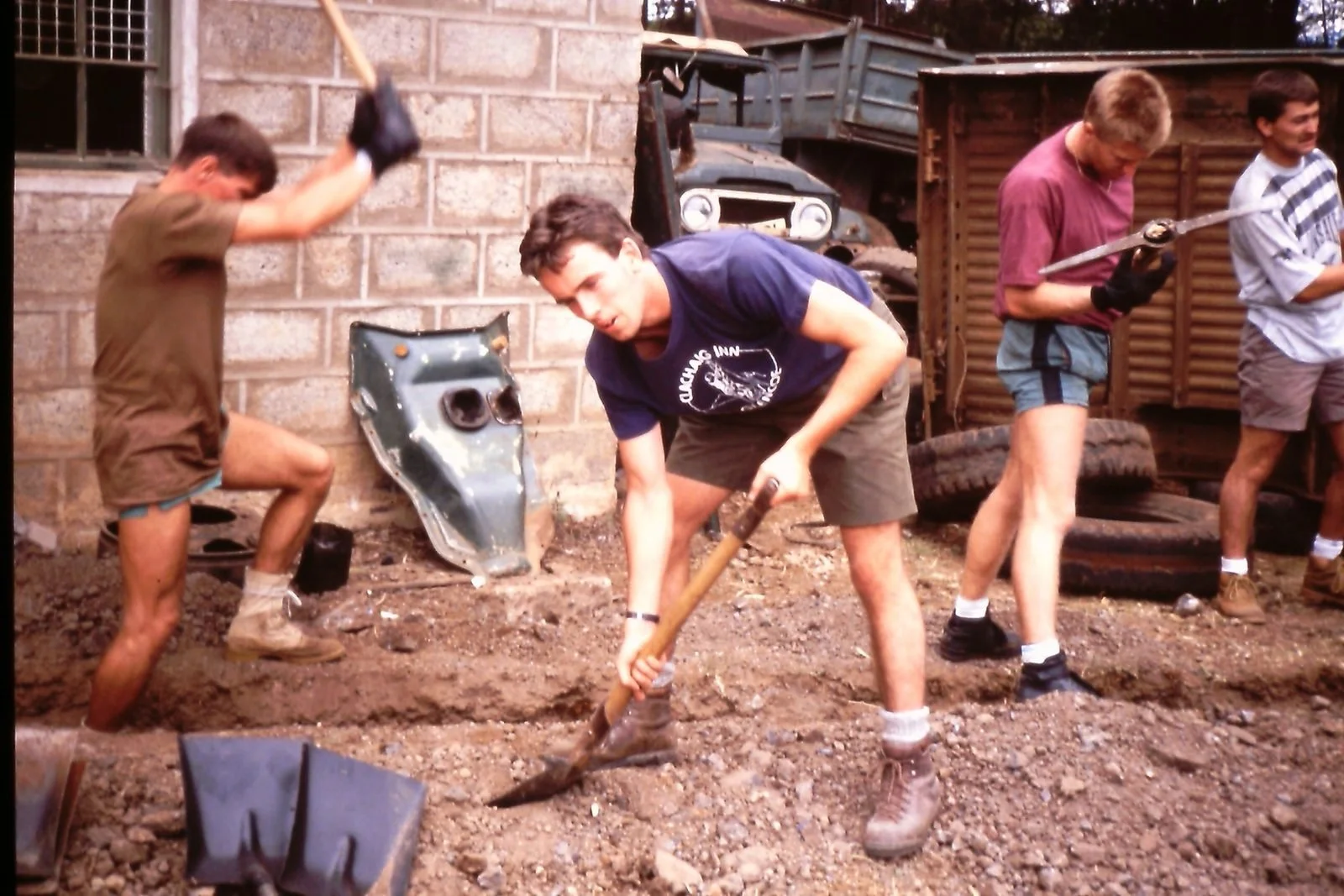 Four young men working outdoors on digging and construction tasks, with brick wall and discarded tires in the background.