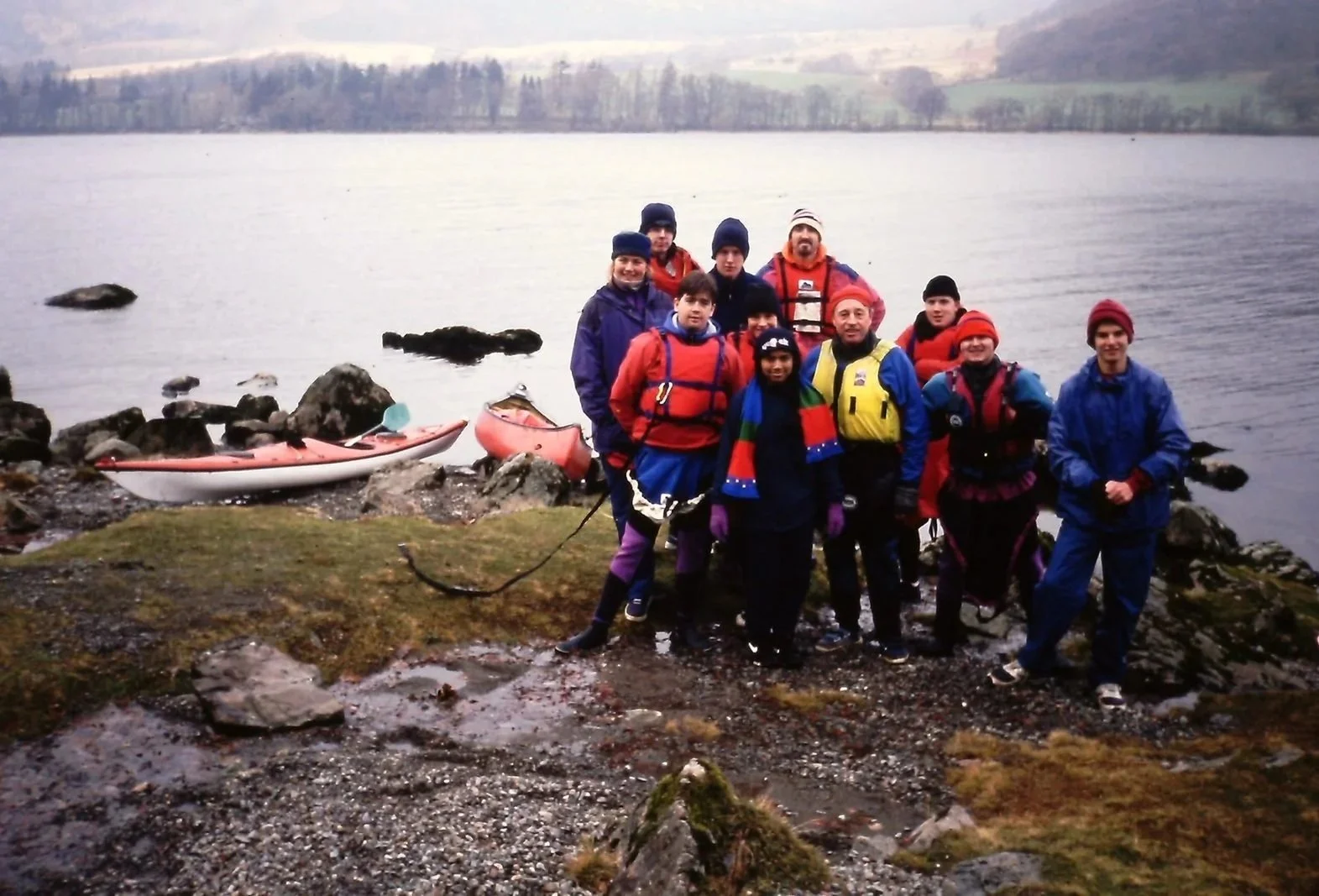 Group of people standing by a lake shore with kayaks, dressed in outdoor gear for kayaking or hiking, with trees and a distant hilly landscape in the background.