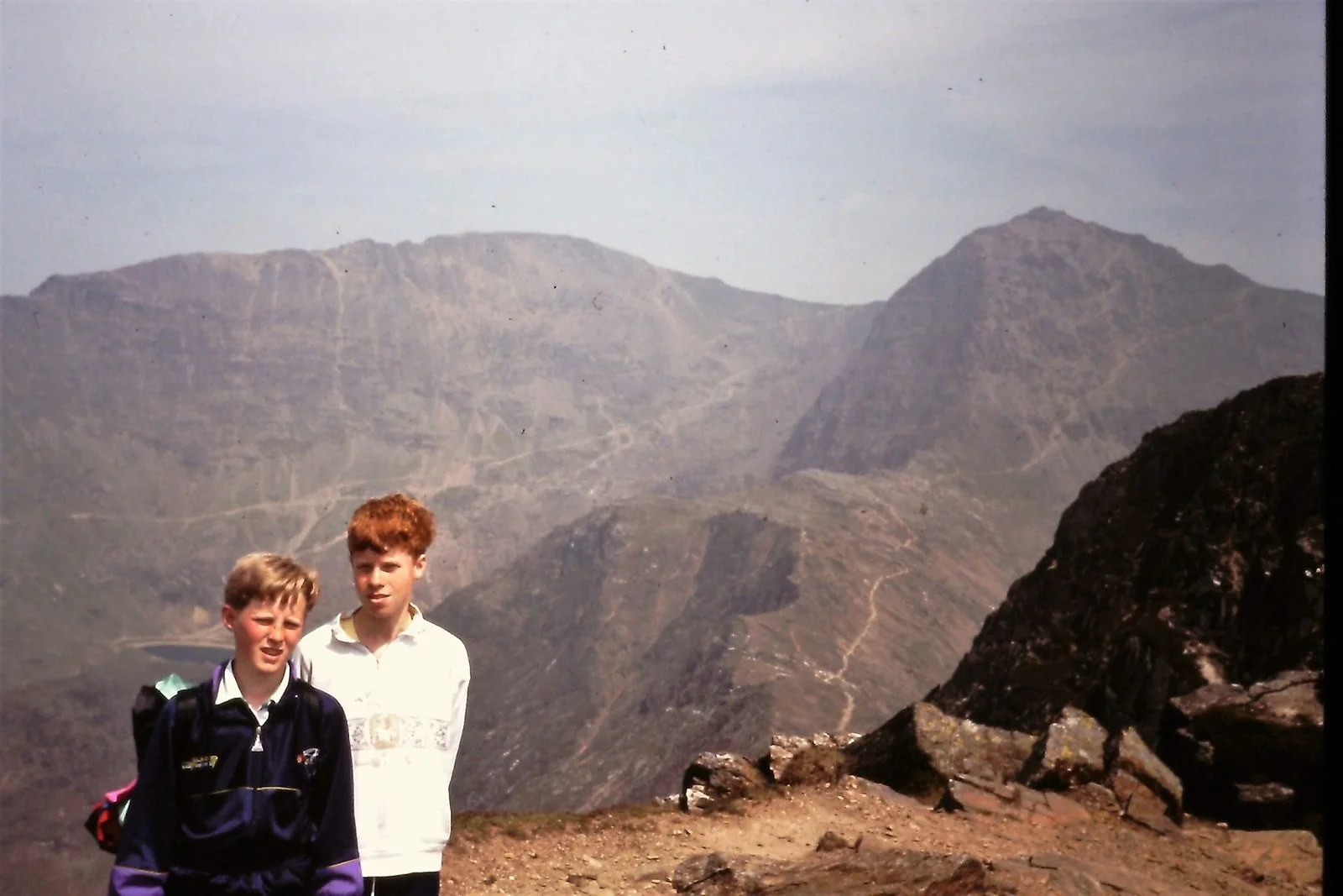 Two boys with backpacks standing on a mountain trail, with rugged mountain peaks in the background.